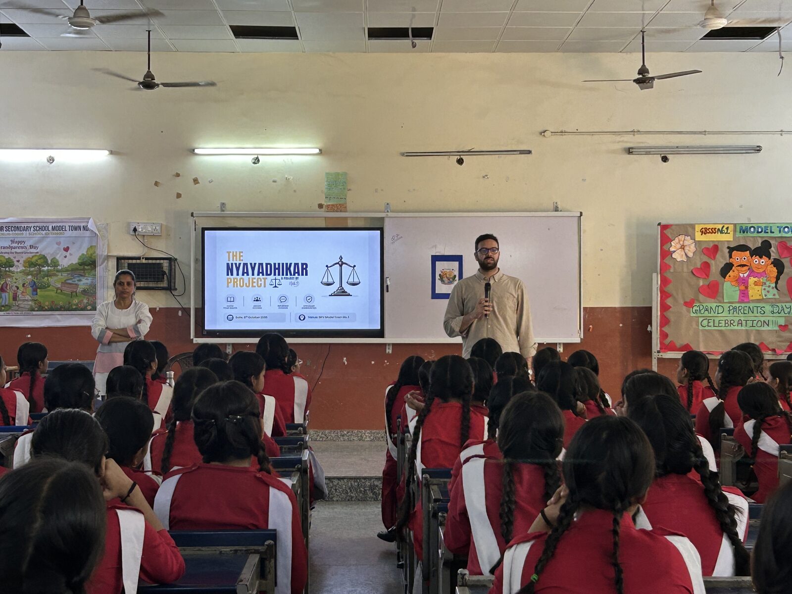 A presenter addresses a room of students standing beside a large display showing a rights awareness project slide.