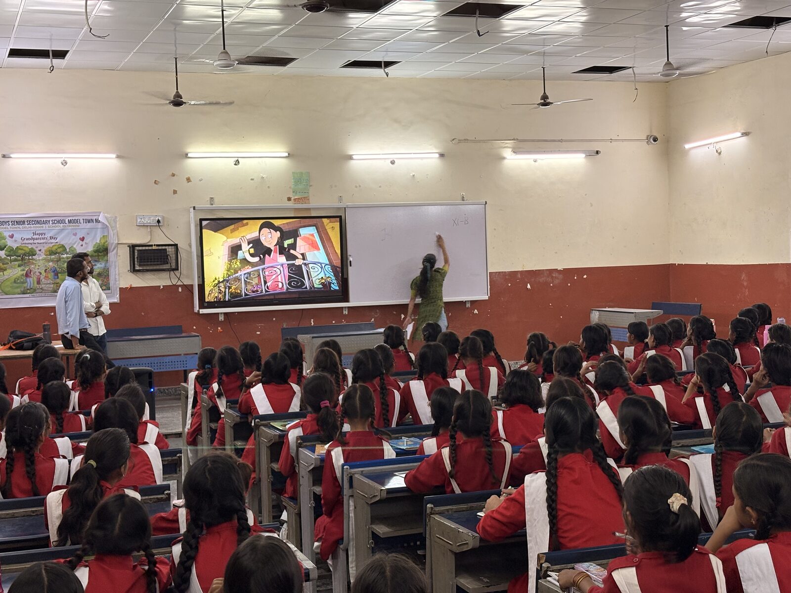 Students in school uniform watching an animated lesson on a classroom display while a facilitator writes on the whiteboard beside them.