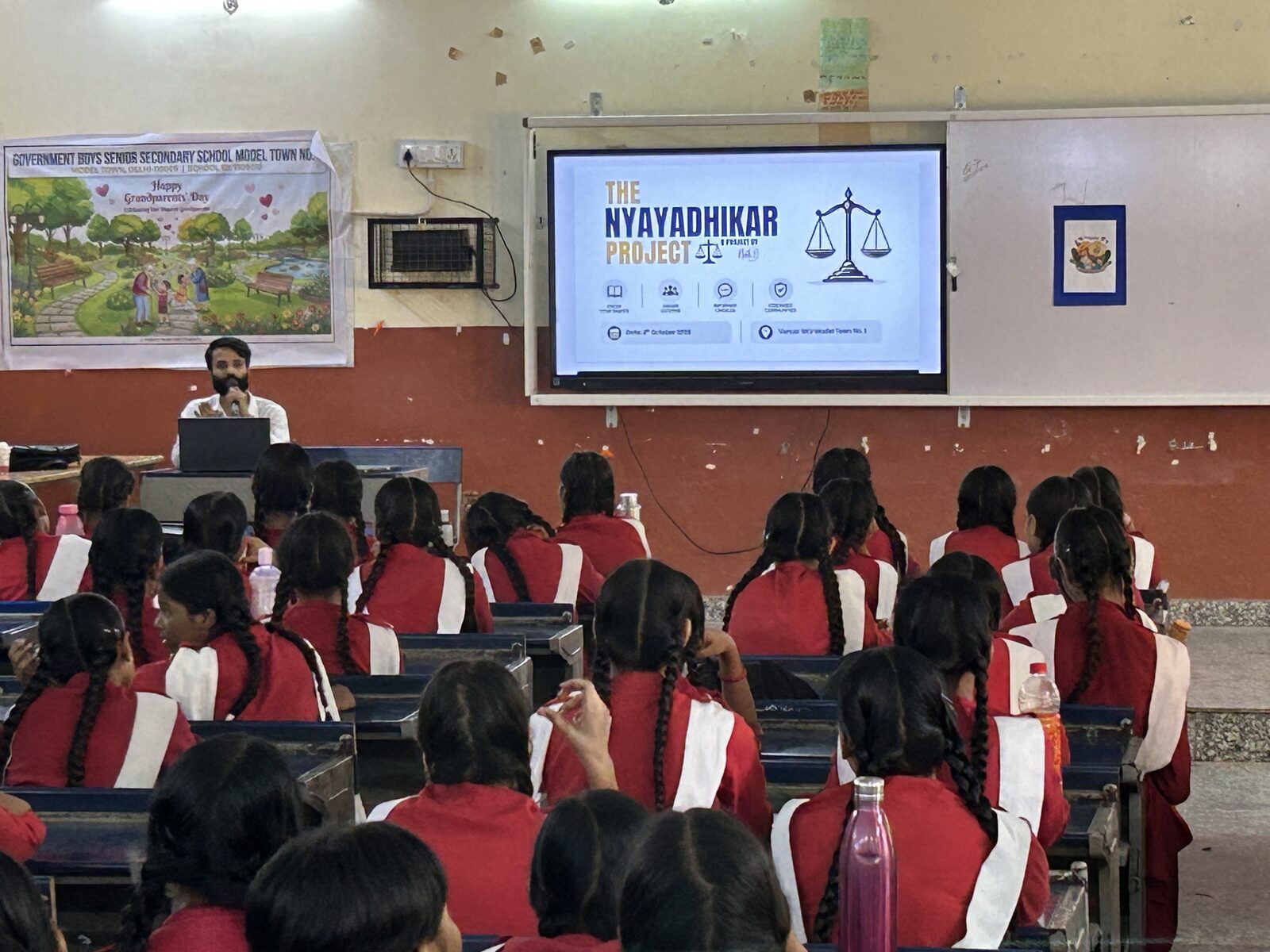 A seated volunteer speaks into a handheld microphone behind an open laptop while rows of students face the classroom display.
